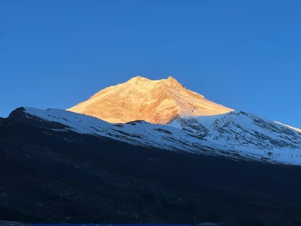A majestic view of Mount Manaslu peak glowing in golden morning sunlight against a clear blue sky, with dark, snow-dusted ridges.
